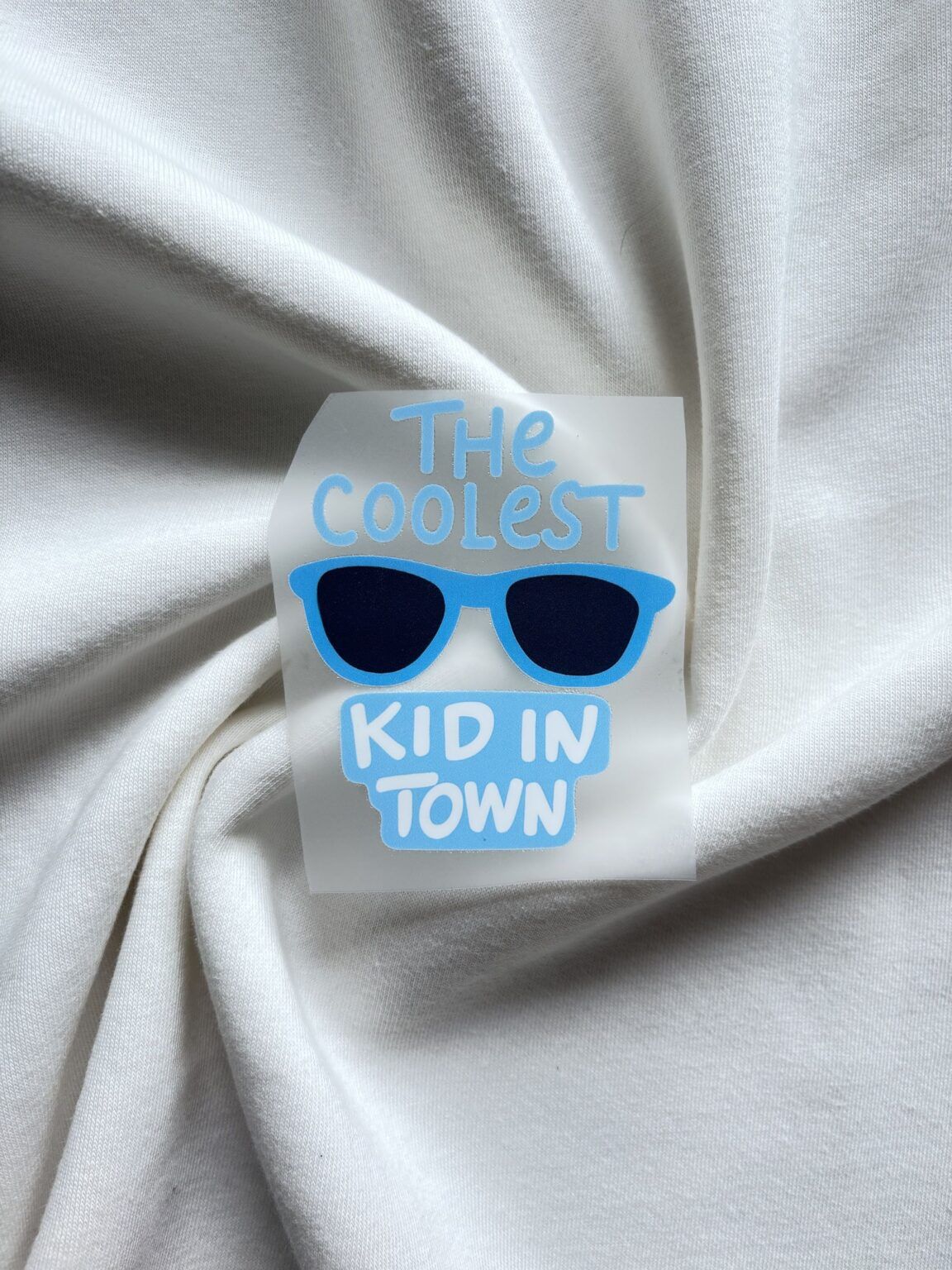 Sticker met tekst The Coolest Kid in Town en blauwe zonnebril op witte katoenen stof